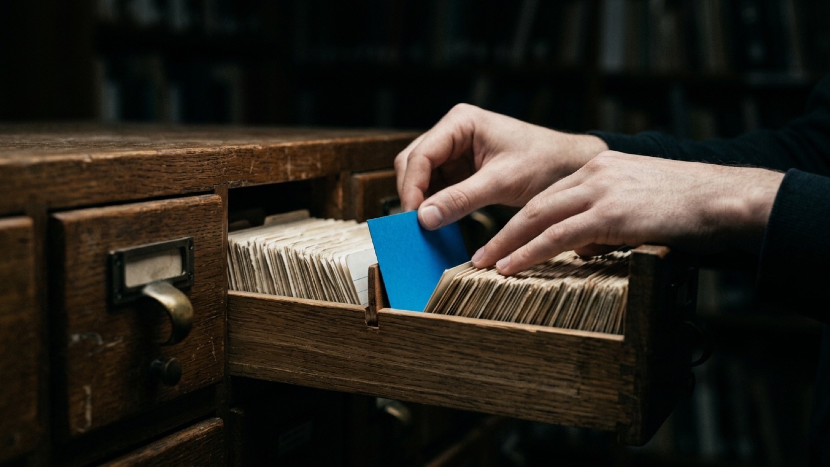 Two hands filing a single blue index card into the open drawer of a wooden card catalog